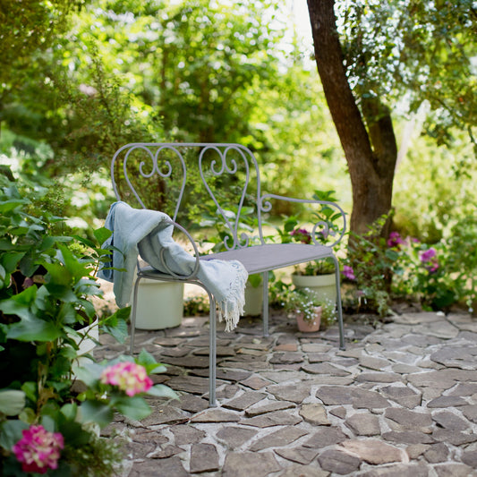 French style metal bench with heart scroll back in a garden setting with cobbled stones and trees and palnts