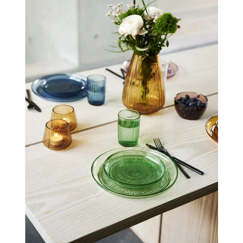 Dining table setting with green glassware, flowers, and cutlery on a light wooden surface.