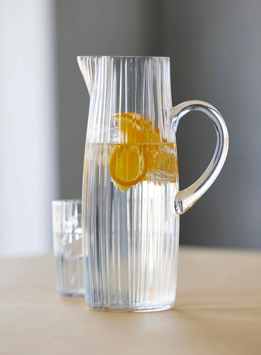 Clear glass pitcher with handle filled with water and an orange slice on a neutral background