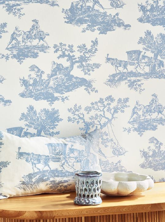 Wallpaper with blue and white toile design featuring horses and carriages, with a pillow and decorative items on a table.