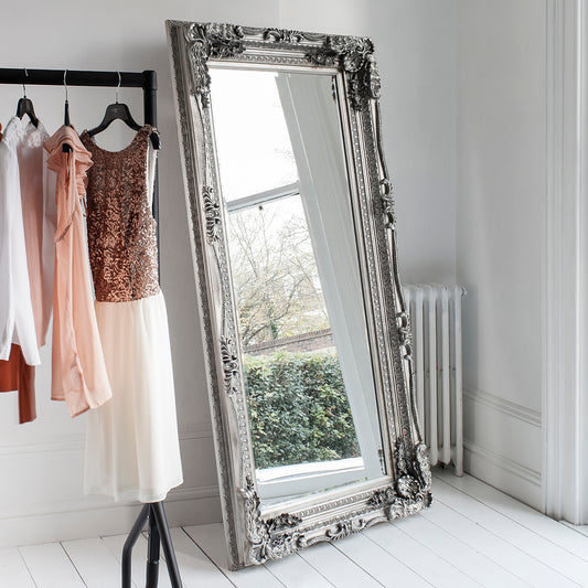 Ornate silver mirror leaning against a white wall in a dressing from with evening wear hanging on a rail
