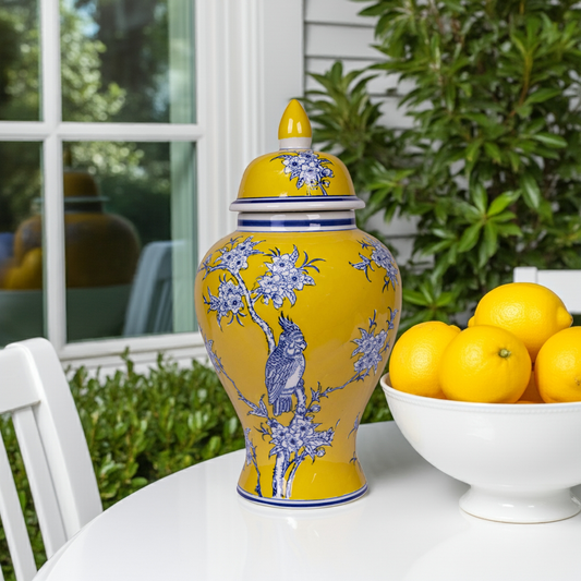 Yellow ceramic vase with blue floral and bird design next to a bowl of lemons on a table outdoors.