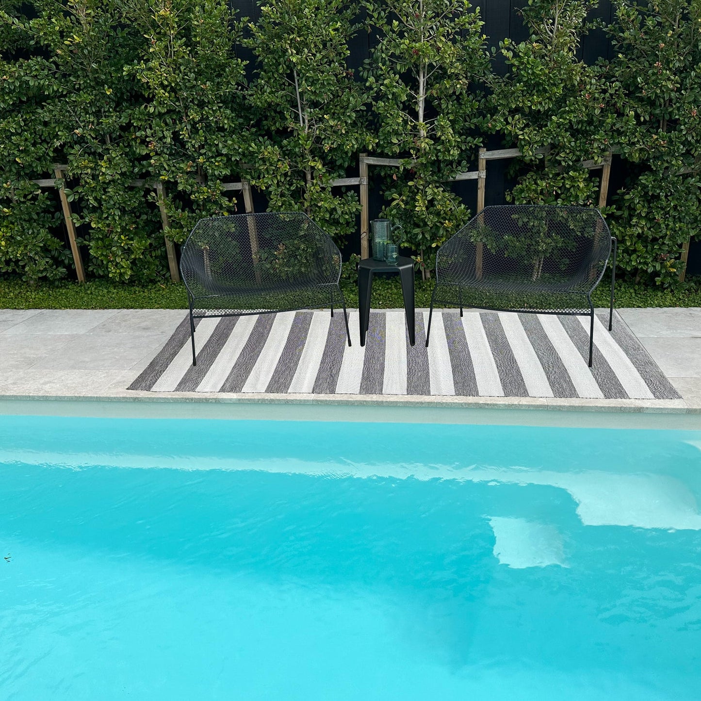 Two black chairs and a small table by a poolside with greenery in the background