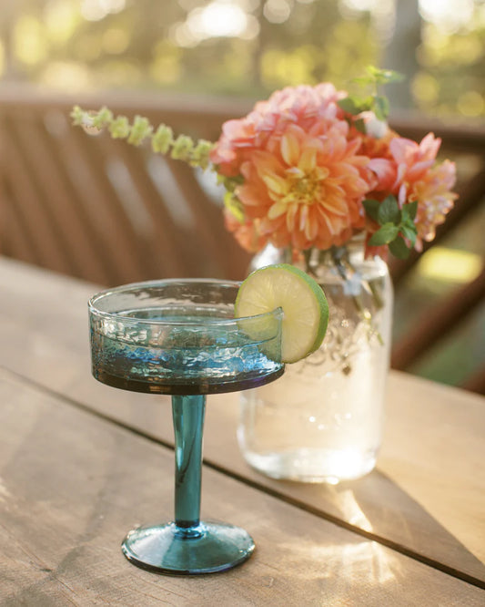 Blue glass cocktail glass with a lime wedge on a wooden table with flowers in the background