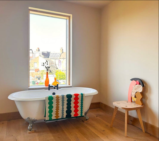 White freestanding bathtub with a colorful towel next to a window in a room with wooden flooring.