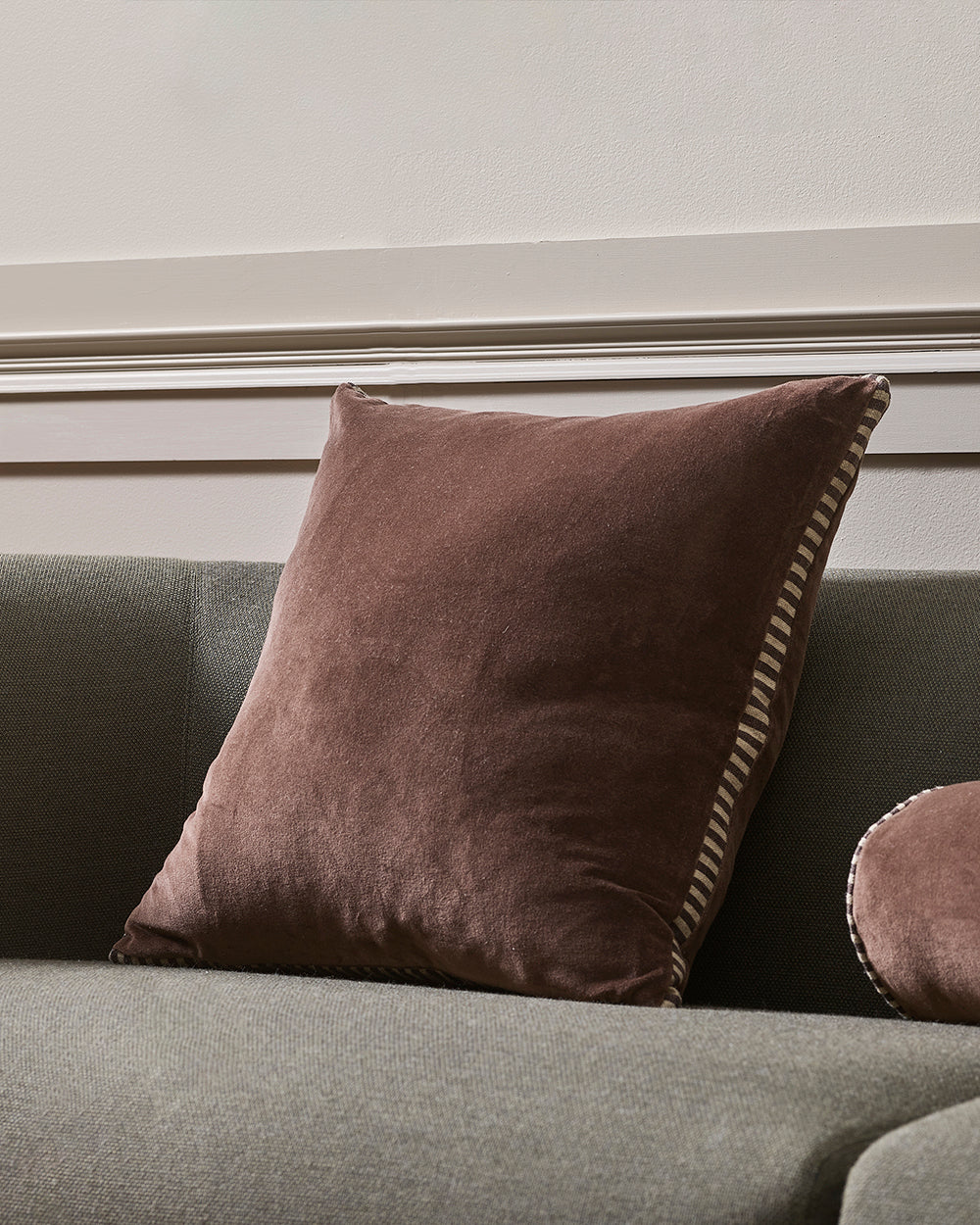 Brown pillow with decorative trim on a gray sofa against a neutral wall.