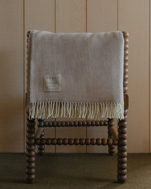 Beige blanket with fringes draped over a wooden chair against a wooden paneled wall.