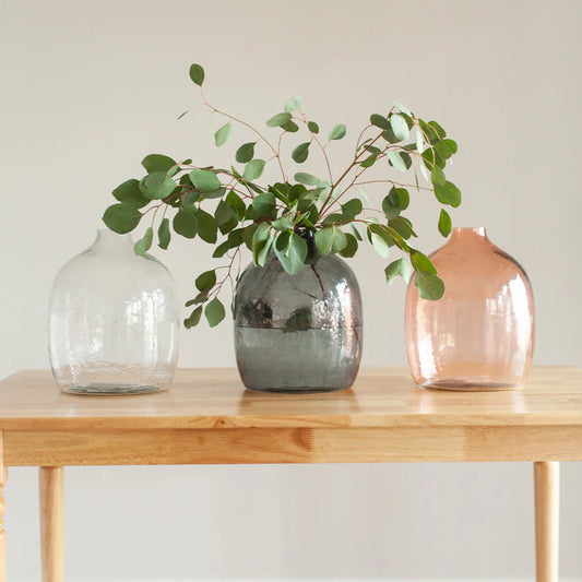Three vases on a wooden table with greenery, one of which is transparent, another gray, and the third pink.
