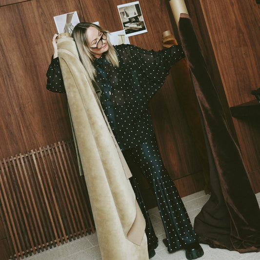 Designer Sarah Ellison hold two rolls of velvet mohair fabric in cream and chocolate. Wearing a polkadot trouser and blouse outfit standing in fron tof a dark wooden wall