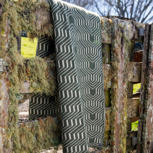 A woven throw blanket with a dark green and cream geometric chevron pattern draped over a rustic, moss-covered wooden fence outdoors.