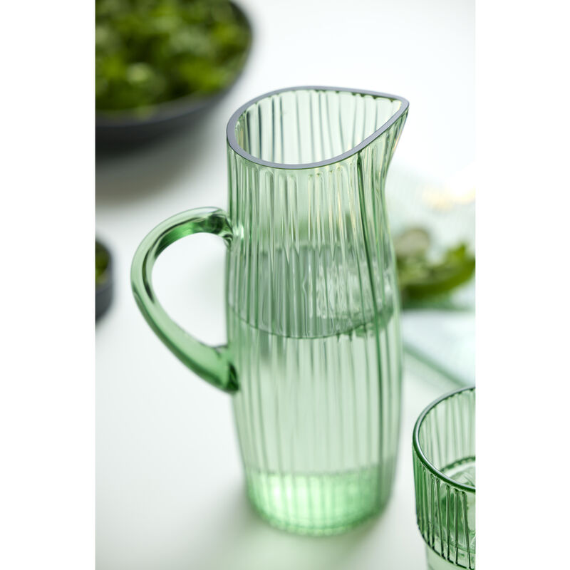 Green glass pitcher on a white surface with blurred greenery in the background