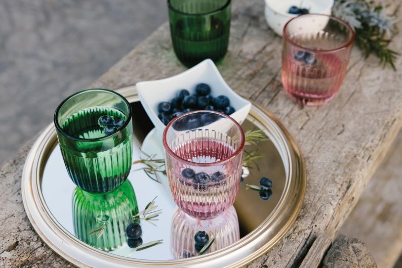 Three ribbed glass tumblers in green, pink, and dark green, arranged on a round mirrored tray with blueberries and rosemary on a rustic wooden table.