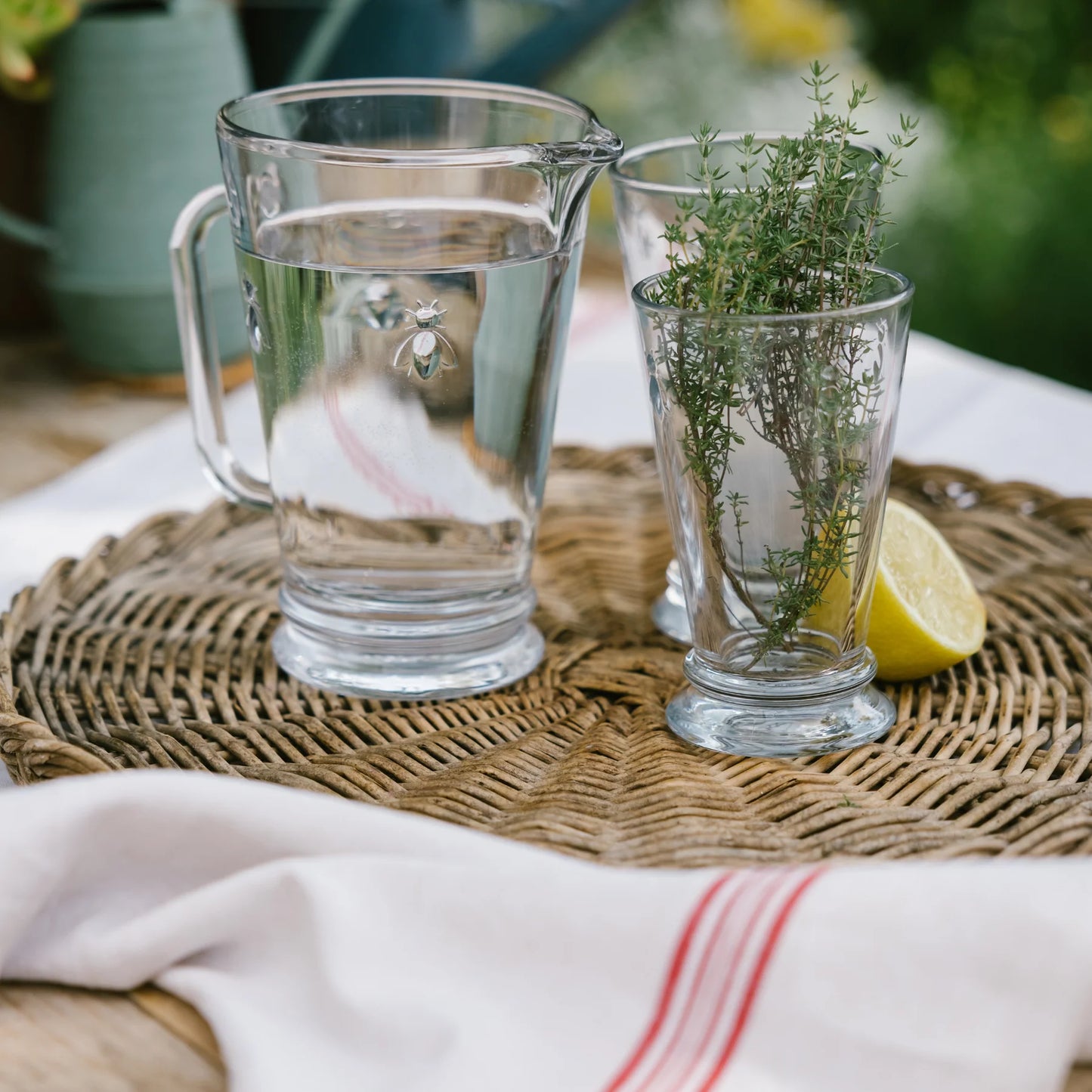 Clear glass pitcher and cups on a woven mat with a lemon and herbs, outdoor setting