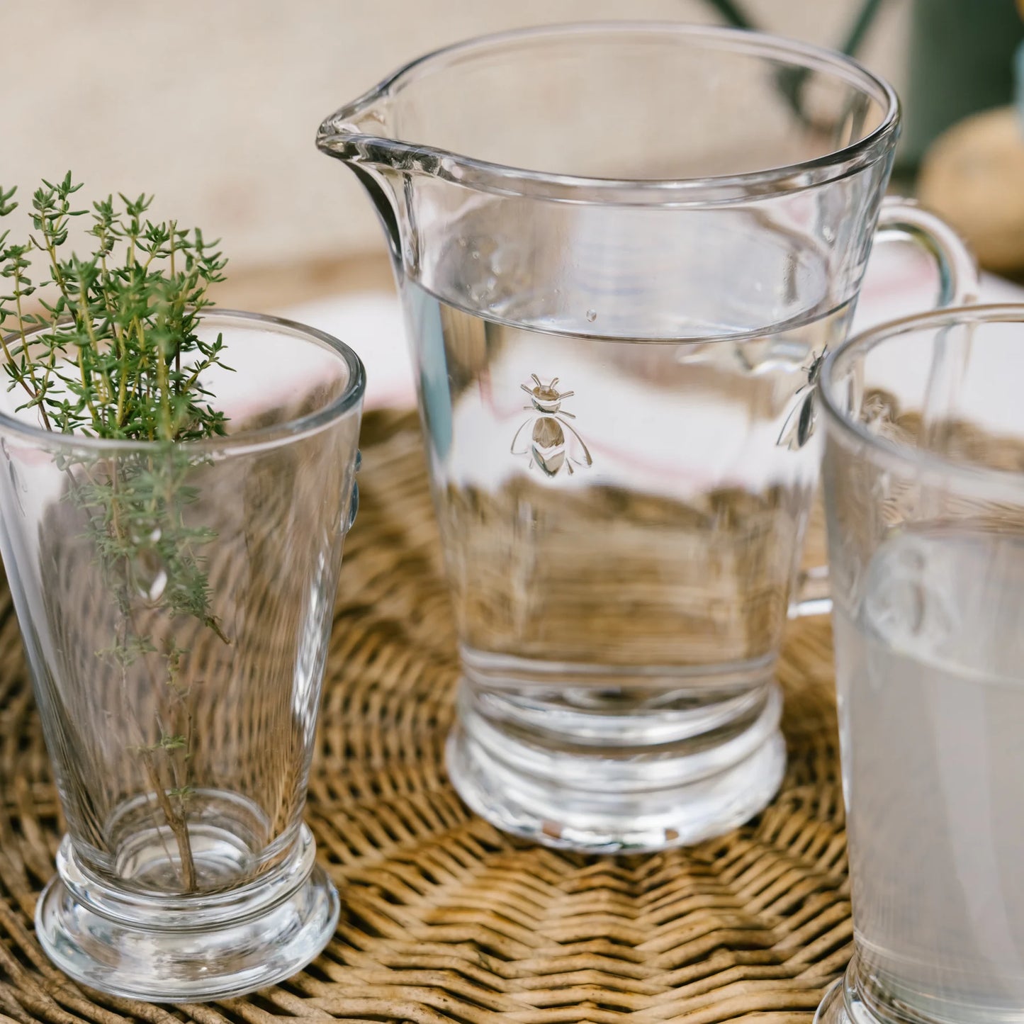 Clear glass pitcher and cups on a woven surface with a plant.