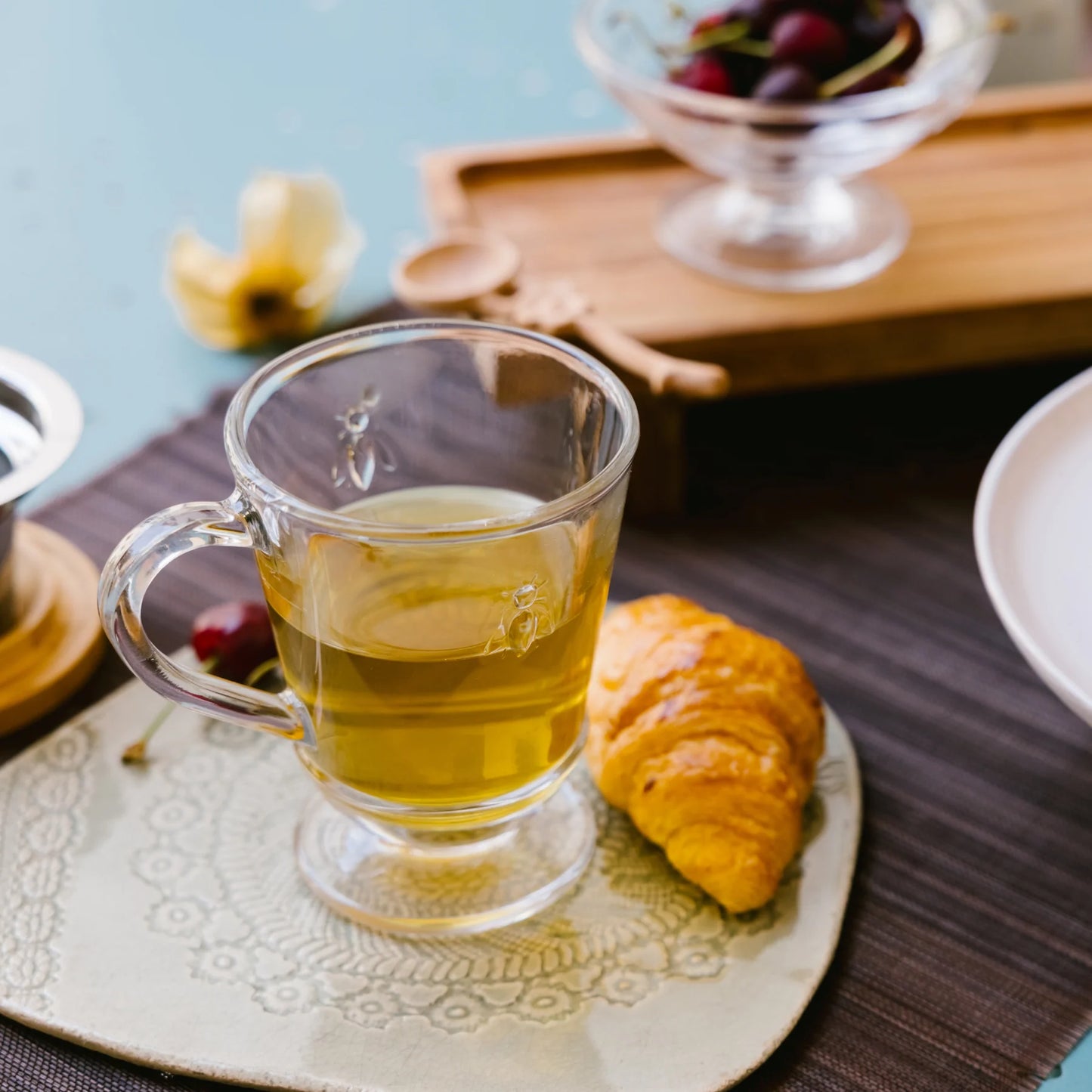 Clear glass mug with green tea on a decorative plate with a croissant and cherry, on a wooden table.