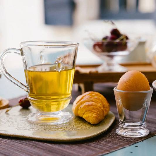 Clear glass mug with yellow liquid, croissant, and egg on a plate with a blurred background