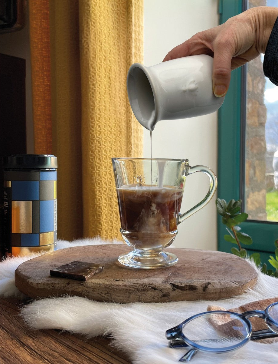 Person pouring coffee from a white mug into a glass mug on a wooden coaster.
