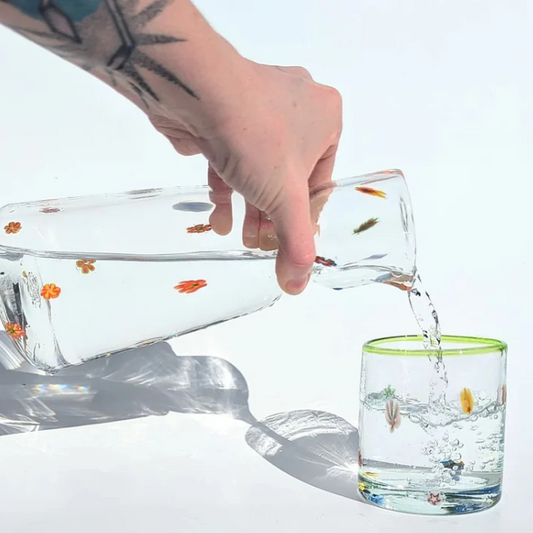 A carafe full of multi coloured flowers sparcely spaced, with water being poured into matching glass