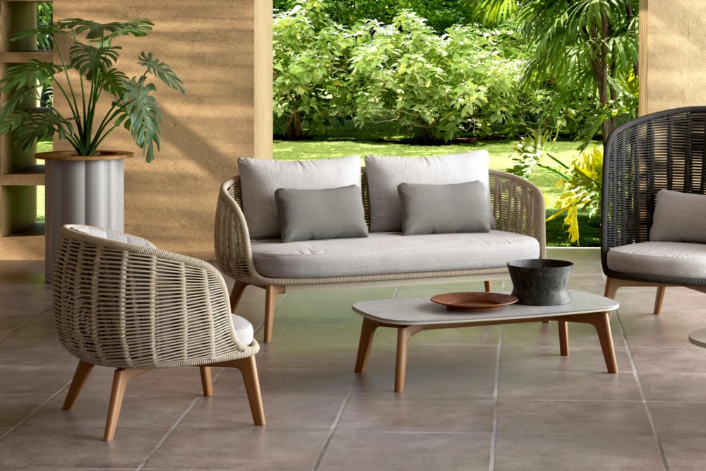 Outdoor patio furniture set with sofa, chairs, and coffee table on a tiled floor with greenery in the background.