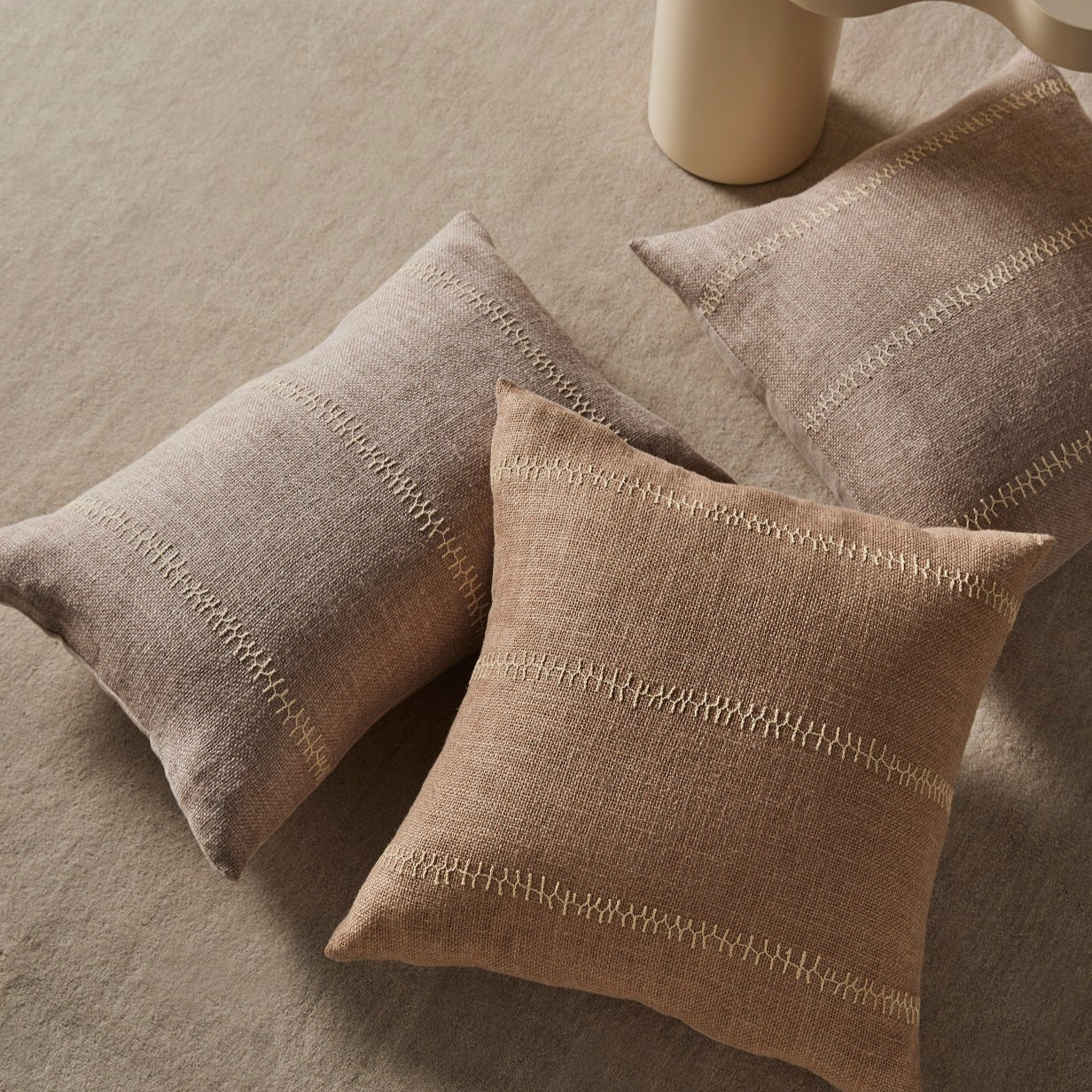 Three beige pillows on a textured surface with a cylindrical object in the background.