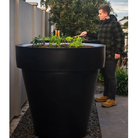 Person standing next to a large black planter with plants on a rooftop garden.