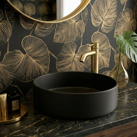 A matte black round vessel sink set into a dark marble countertop, paired with a brushed gold faucet. The background features a matching black and gold botanical wallpaper, with a gold-framed mirror and a green monstera leaf in a glass vase.