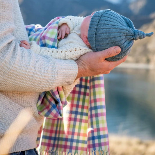 Baby in blue knitted beanie being held by a man and wrapped in a multi check merino baby blanket