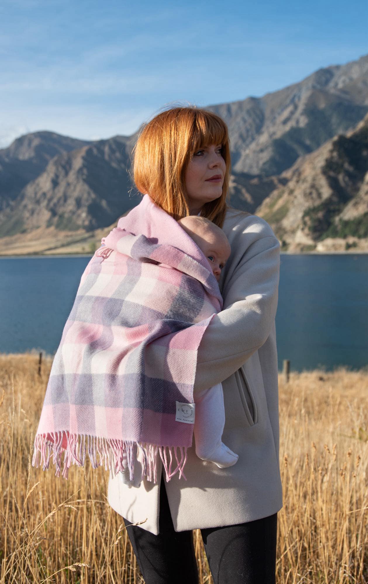 Mother with red hair holding a new born wrapped in a pink and white checked merino blanket with lake and mountains in background