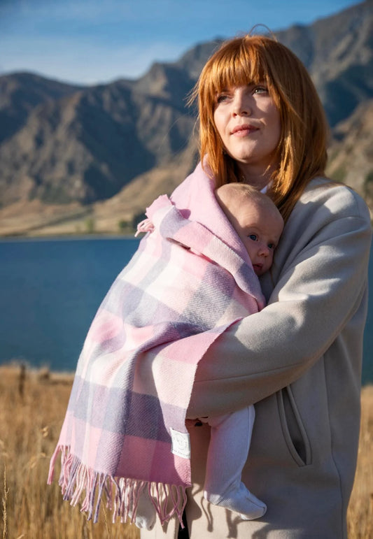 Mother with red hair holding a new born wrapped in a pink and white checked merino blanket with lake and mountains in background