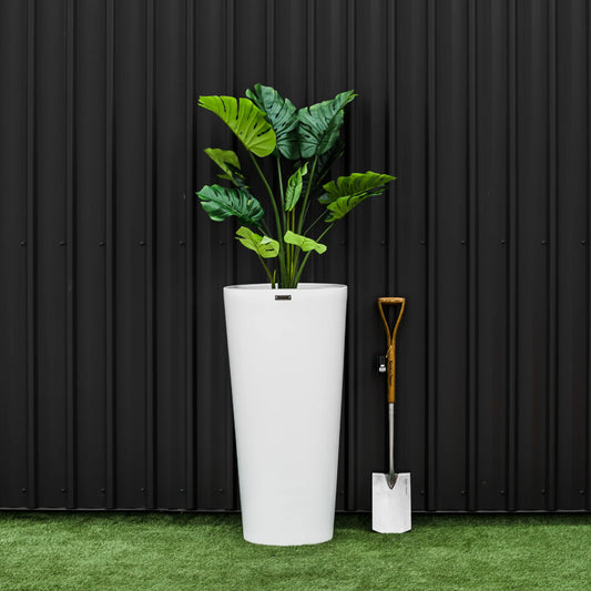 Tall white planter with green plant against a black corrugated metal wall.