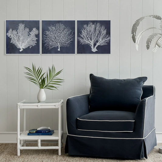 3 art prints with white coral images on a navy background in a room setting with a navy armchair with white piping and a white bamboo coffee table alongside. The walls are white shiplap and a white metal leaf lighting fitting peeps in from the side