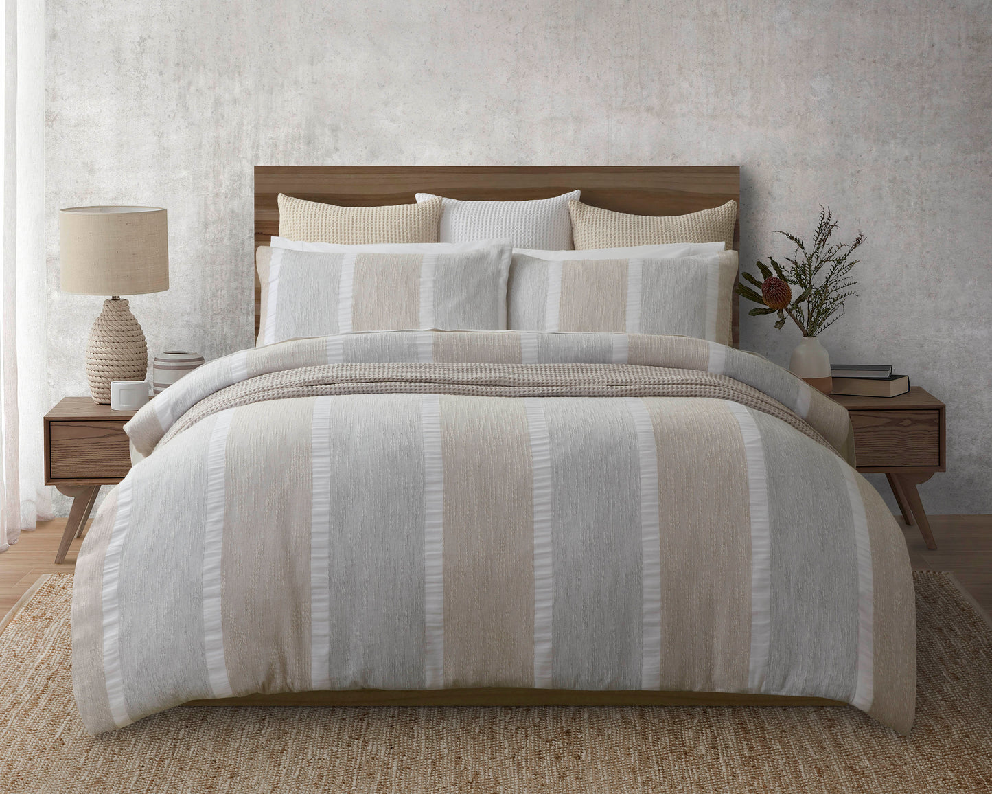 The Nina duvet cover is beige and white colour block striped duvet cover set on a bed with two wooden side tables, one with a lamp and ceramics and the other with a ceramic vase with dried plant lifef and books. Bed sits on a jute rug.