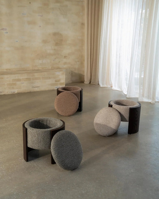 Four modern pouf stools with dark wood frames and textured spherical seat cushions in gray, beige, and taupe, arranged on a concrete floor in a minimalist room with sheer curtains.