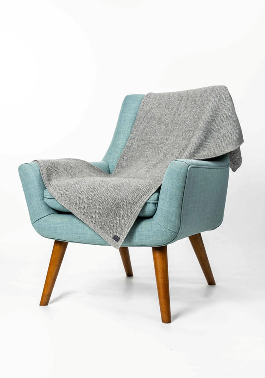 A mid-century modern armchair upholstered in light blue fabric with tapered wooden legs, draped with a heather gray knit throw blanket, set against a plain white studio background.