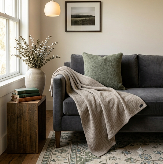 A cozy living room corner featuring a dark gray fabric sofa with a textured green pillow and a beige throw blanket draped over the arm. A rustic wooden side table next to the window holds a vase of dried flowers and books, with a light wood ceiling and a framed landscape print on the wall.