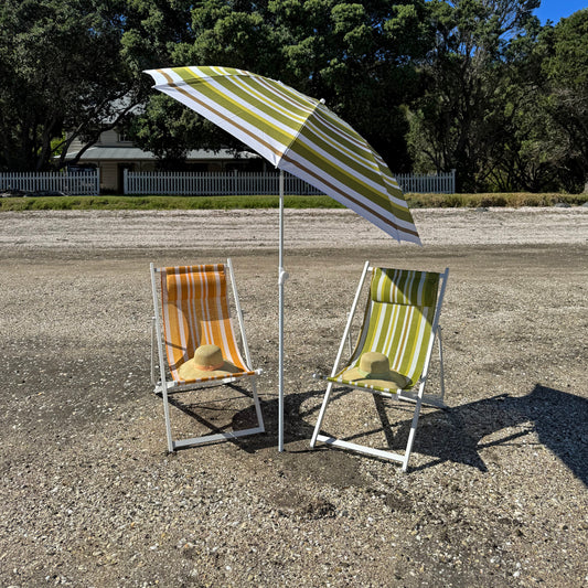 Two beach chairs under a striped umbrella on a gravel surface with trees in the background.