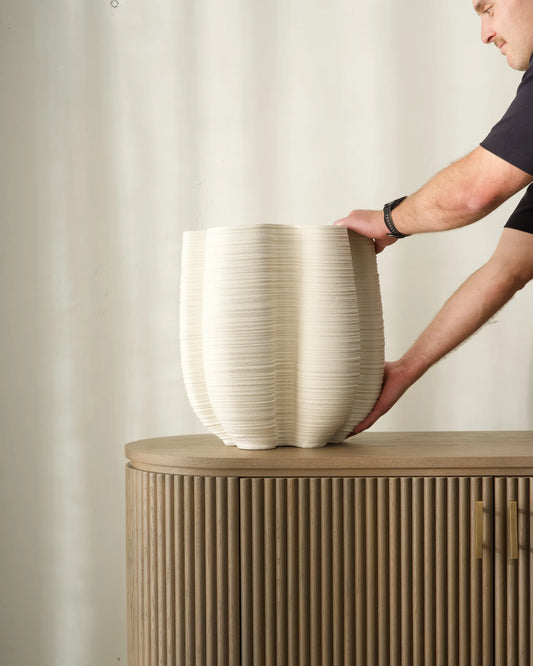 Person placing a large white vase on a wooden cabinet against a plain wall.