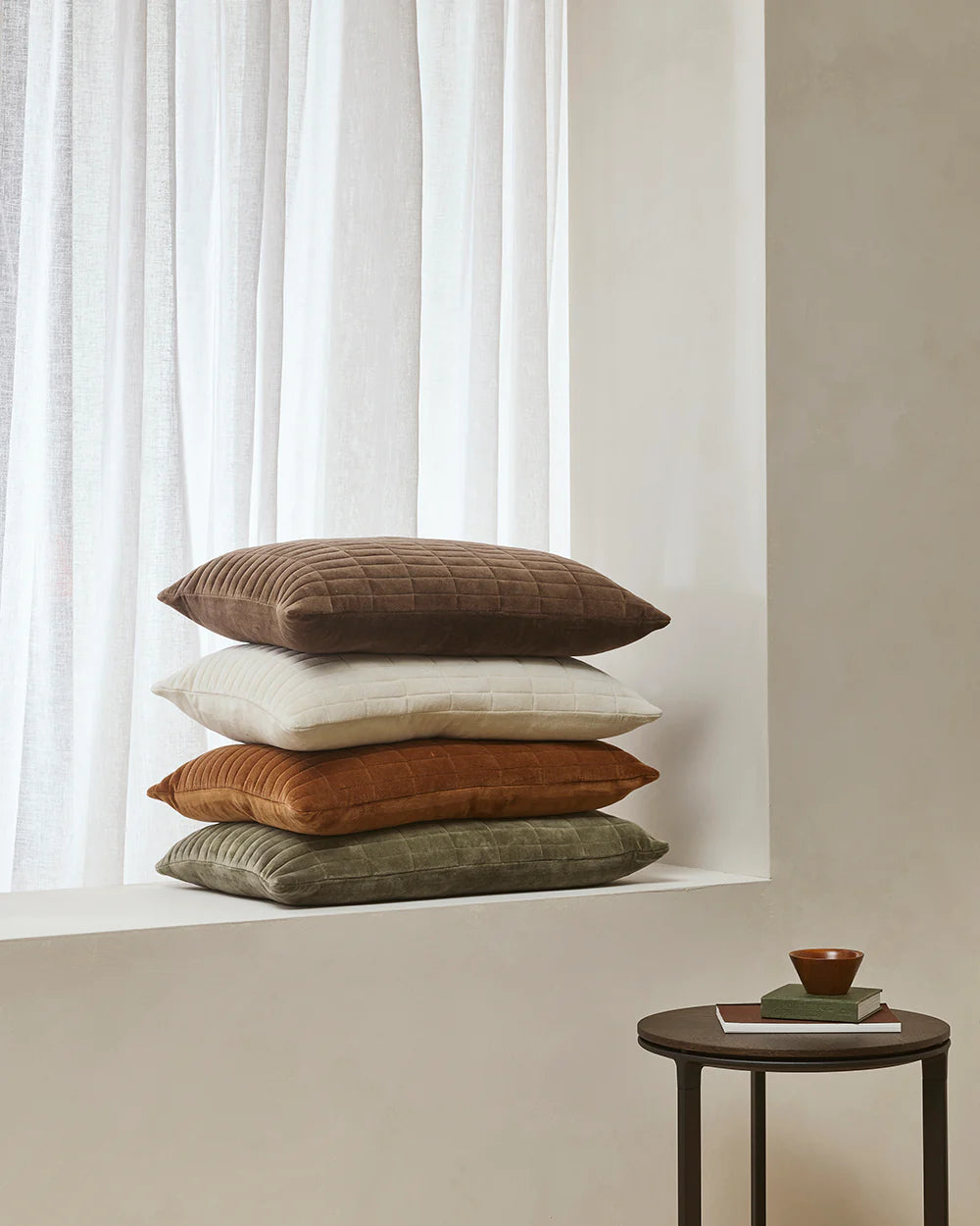 Stack of brown, white, and green cushions on a windowsill with a small table and bowl in the foreground.