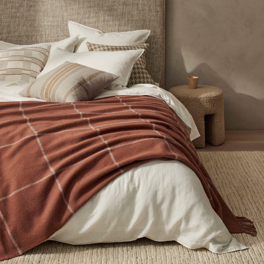 warm toned ranfurly throw with large scale check draped over a bed with white linen and beige walls and head board. Sitting on a pale cream wool carpet with neural colours on the beds cushion. Along sie is a small boucle fabric side table