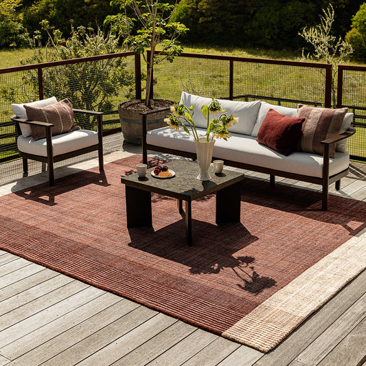 Outdoor patio furniture set on a wooden deck with a rust-red woven area rug, a white sofa with white and red cushions, a matching armchair, and a dark coffee table, with a lush green forest background.