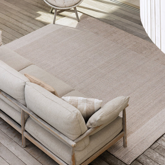 A large rectangular outdoor area rug in a light beige tone, shown on a wooden deck beside a beige sofa and a woven accent chair.