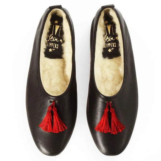 A pair of black leather ballet flats with red tassels and a cream-colored shearling lining, shown from a top-down view on a white background.