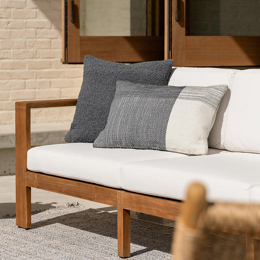Wooden outdoor sofa with white cushions and gray patterned pillows against a brick wall.