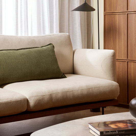 Long dark green cushion on a moderl cream sofa with wood paneling and a black floor lamp to the right and a white marble coffee table stacked with books in front of sofa