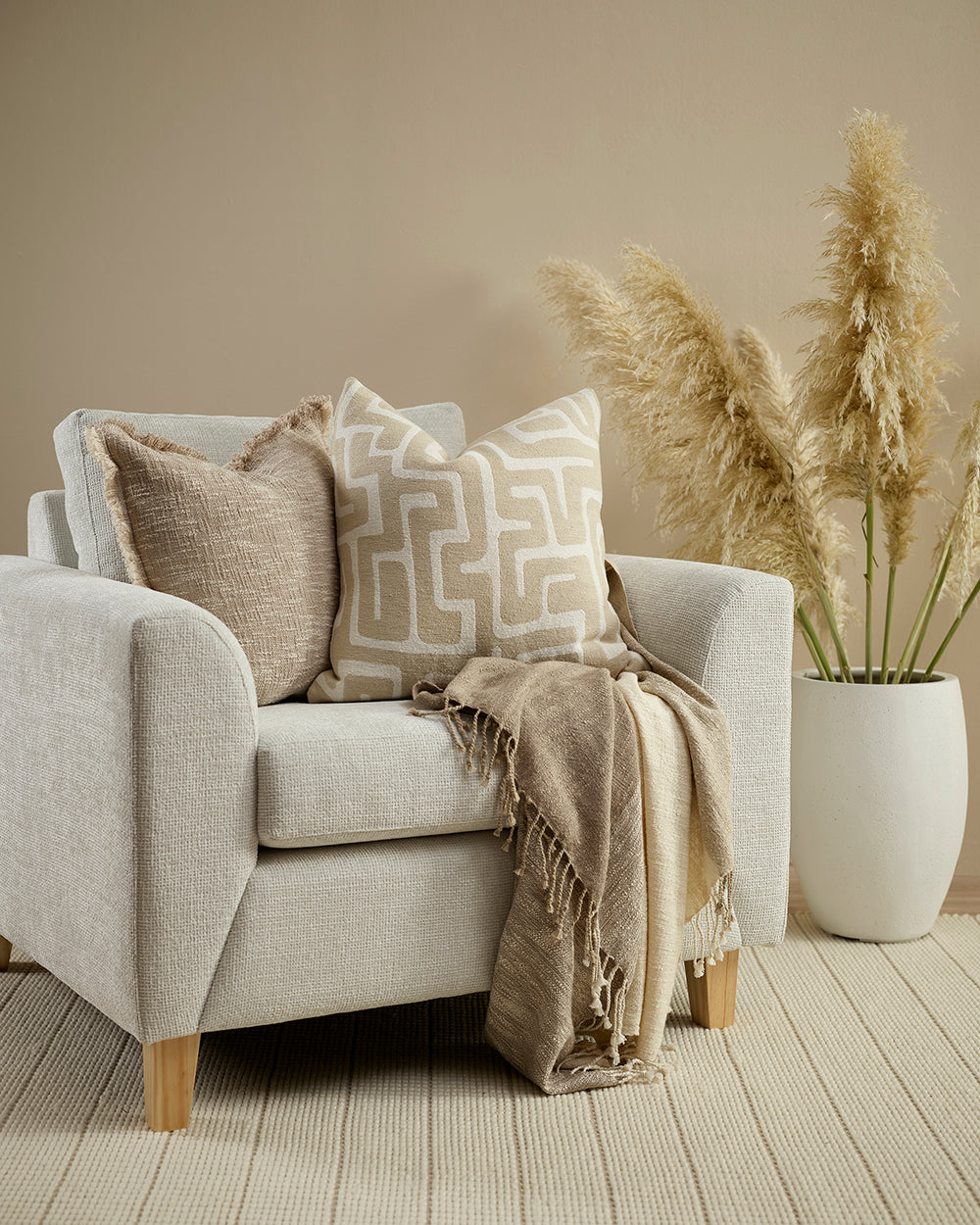 Beige armchair with patterned cushions and a blanket, accompanied by a vase with dried plants on a wooden floor.