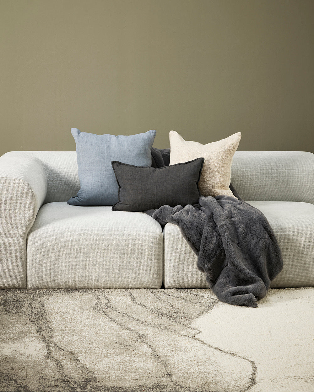 Cloud blue, ecru and charcoal cushions sitting on a whitel modern sofa with sage green walls and cream patterned rug