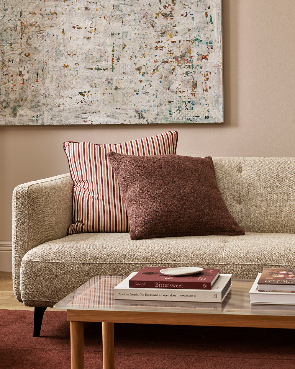 A beige textured sofa in a living room setting, styled with two throw pillows: one solid dark brown and one red and white striped. A glass-top coffee table with books sits in front, and a large abstract painting hangs on the beige wall behind.