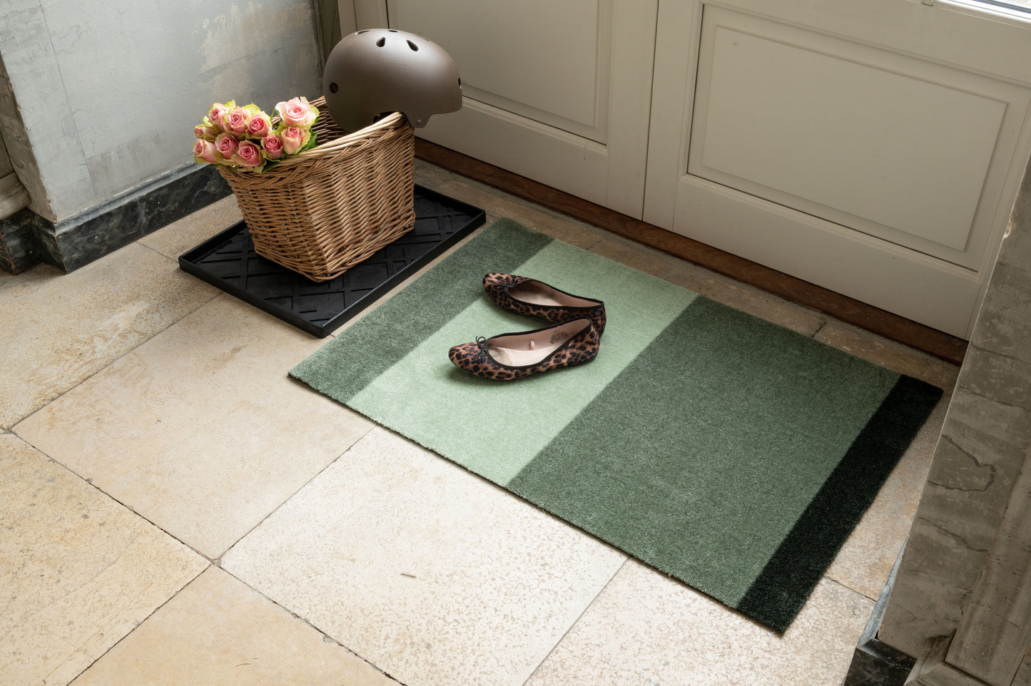 Green doormat with a pair of shoes on a tiled floor next to a basket with flowers and a helmet.