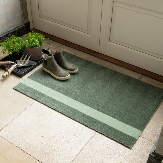 Green doormat with striped pattern on a tiled floor, next to a pair of boots and gardening tools.