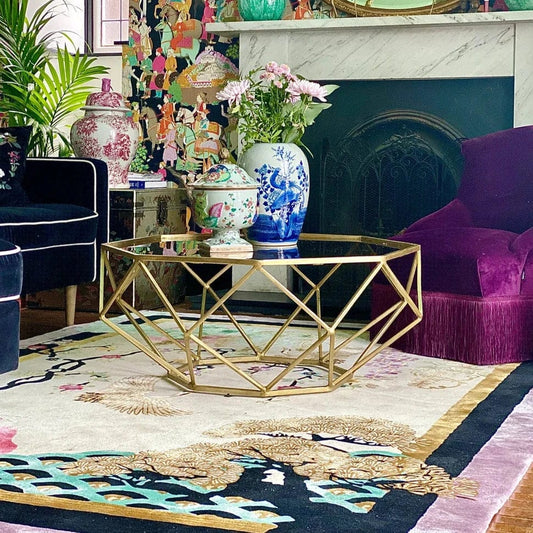 Wendy Morrison rug Eternity Light covereind in chinoiserie symbols like flowers and cranes, surrounded by opulent furniture, gold geometric caged coffee table with chiness vases on top, and a navy and a purple velvet seating around it. A white marble fireplace is the background with plants and more chinese style urns dotted around